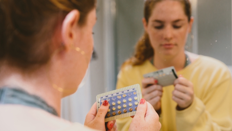 A woman looks at a pack of birth control pills in the mirror